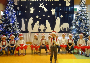 Chłopiec recytuje wiersz, trzyma w ręku mikrofon w tle dzieci siedzą na krzesłach na tle dekoracji świątecznej, choinek.